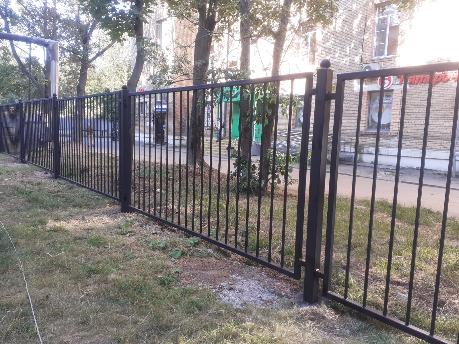 Чёрный металлический забор с вертикальным заполнением вдоль озеленённой территории у здания
