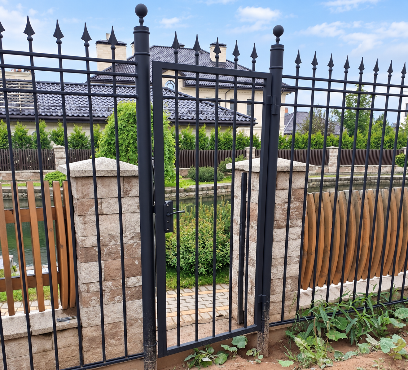 Чёрная калитка с прямыми вертикальными прутьями и пиками на круглых металлических столбах у загородного дома